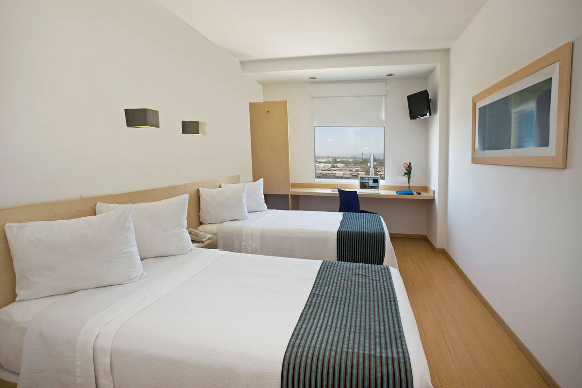 Habitació d'hotel amb dos llits, una taula de treball, un televisor i una finestra amb vista a la ciutat.