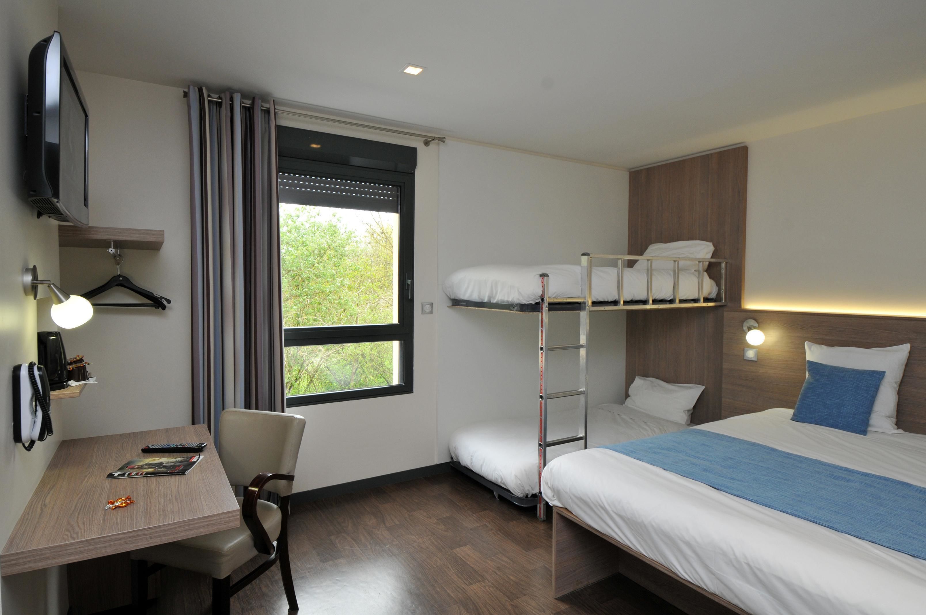 Une chambre d’hôtel avec un lit superposé, un bureau avec une chaise, un téléviseur fixé au mur et une fenêtre avec des rideaux.