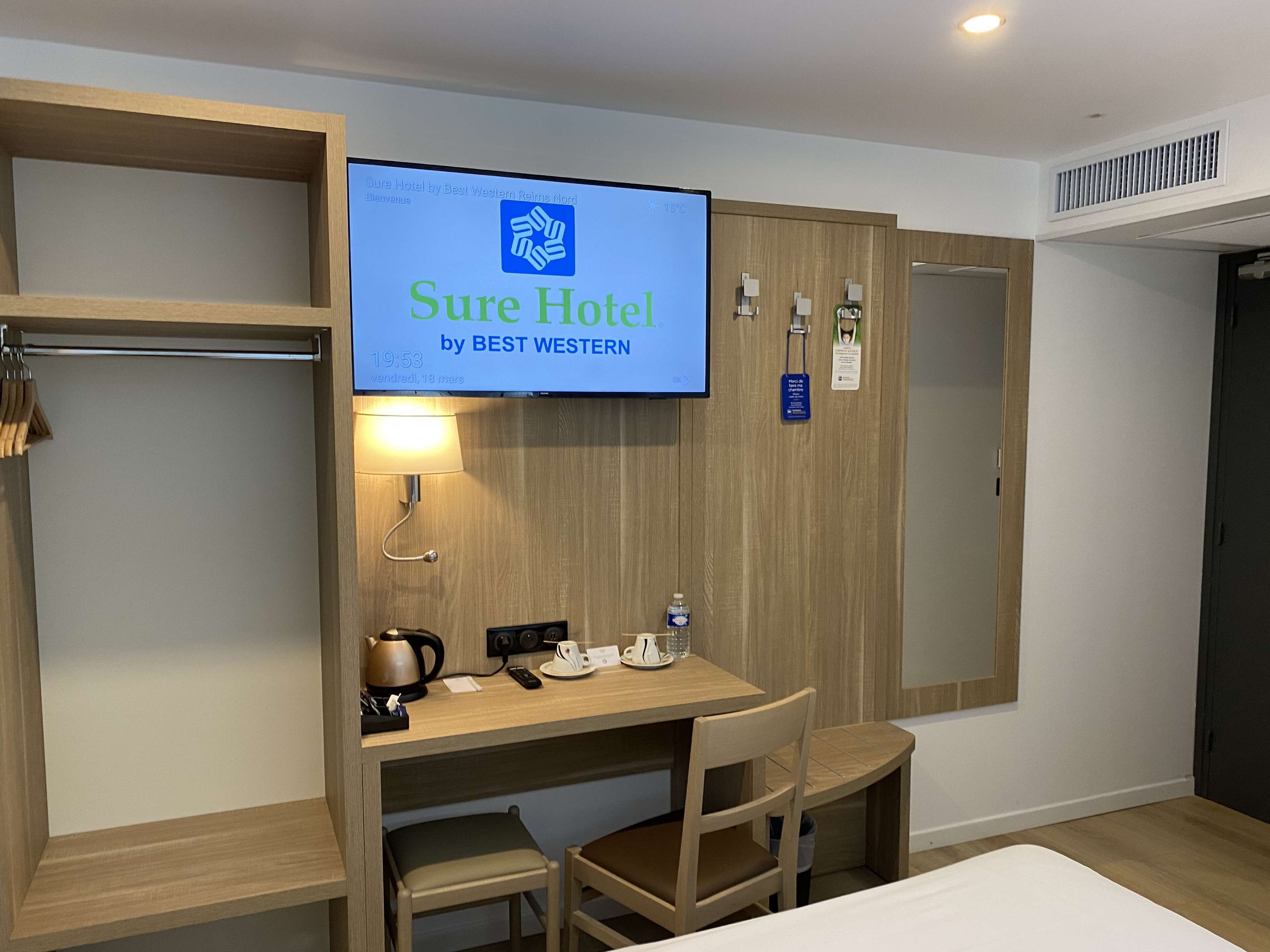 Une chambre d’hôtel équipée d’un lit, d’un bureau avec une bouilloire, des tasses et un téléphone, ainsi que d’un téléviseur affichant le nom de l’hôtel.