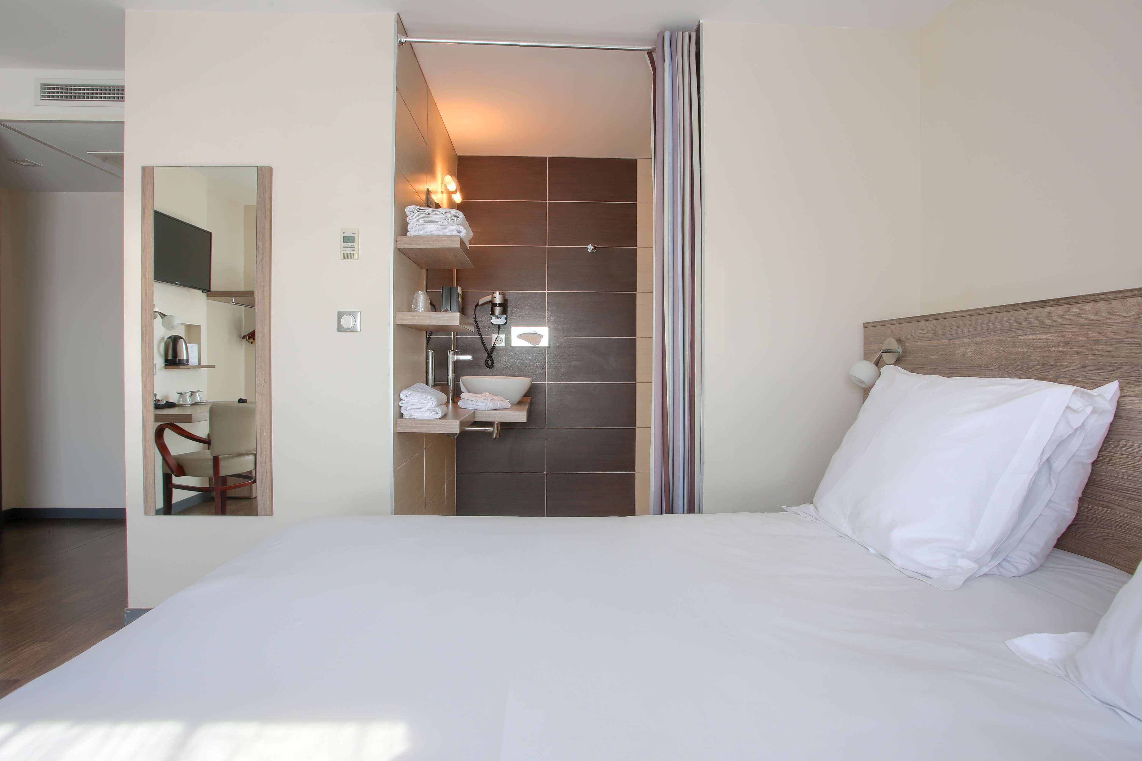 Quarto de hotel com uma cama grande, cabeceira de madeira, banheiro com chuveiro, espelho e prateleira com toalhas e amenities.