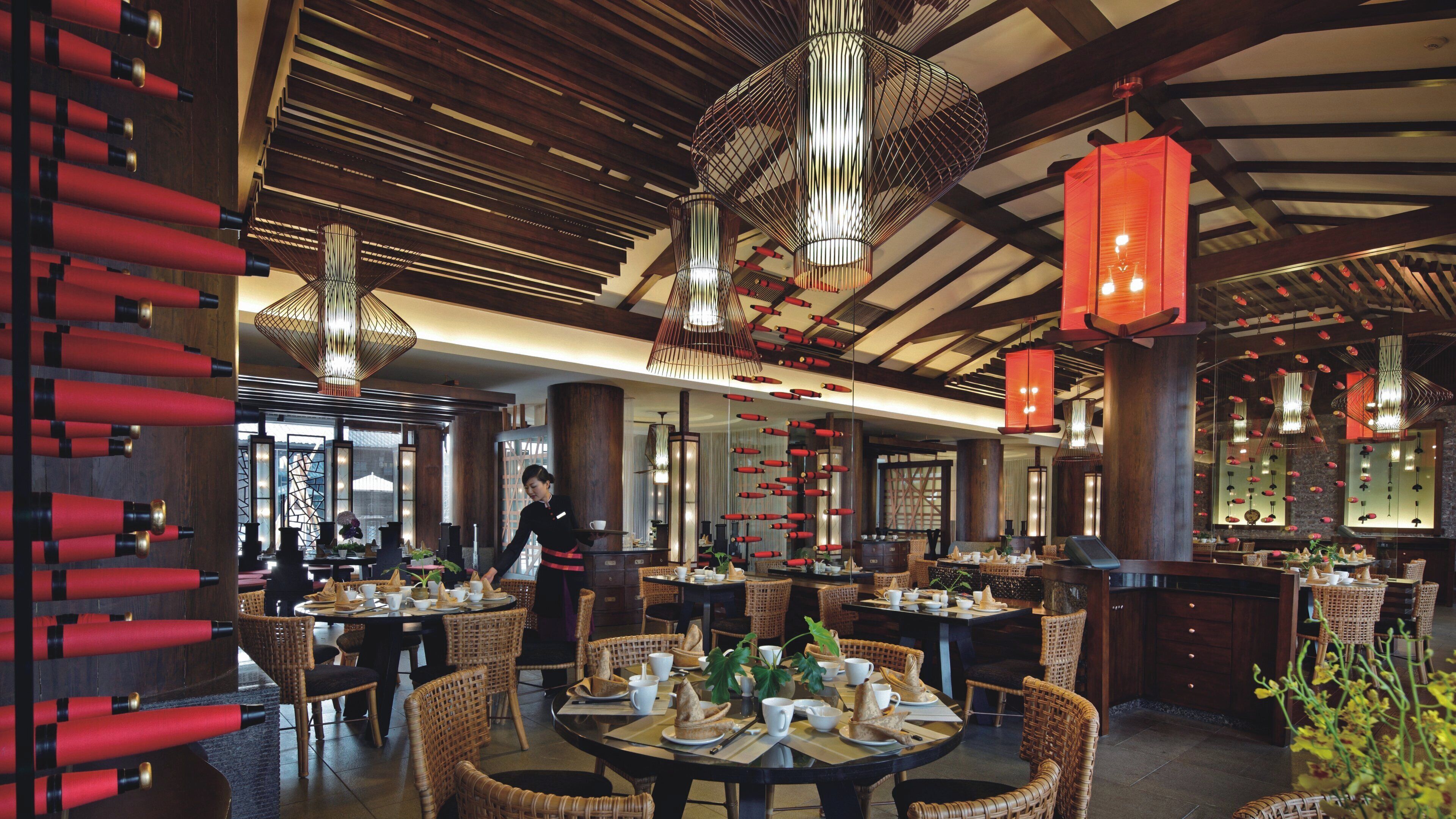 A restaurant interior with high ceilings, exposed beams, and modern pendant lighting. Tables are set with white tableware and wicker chairs.