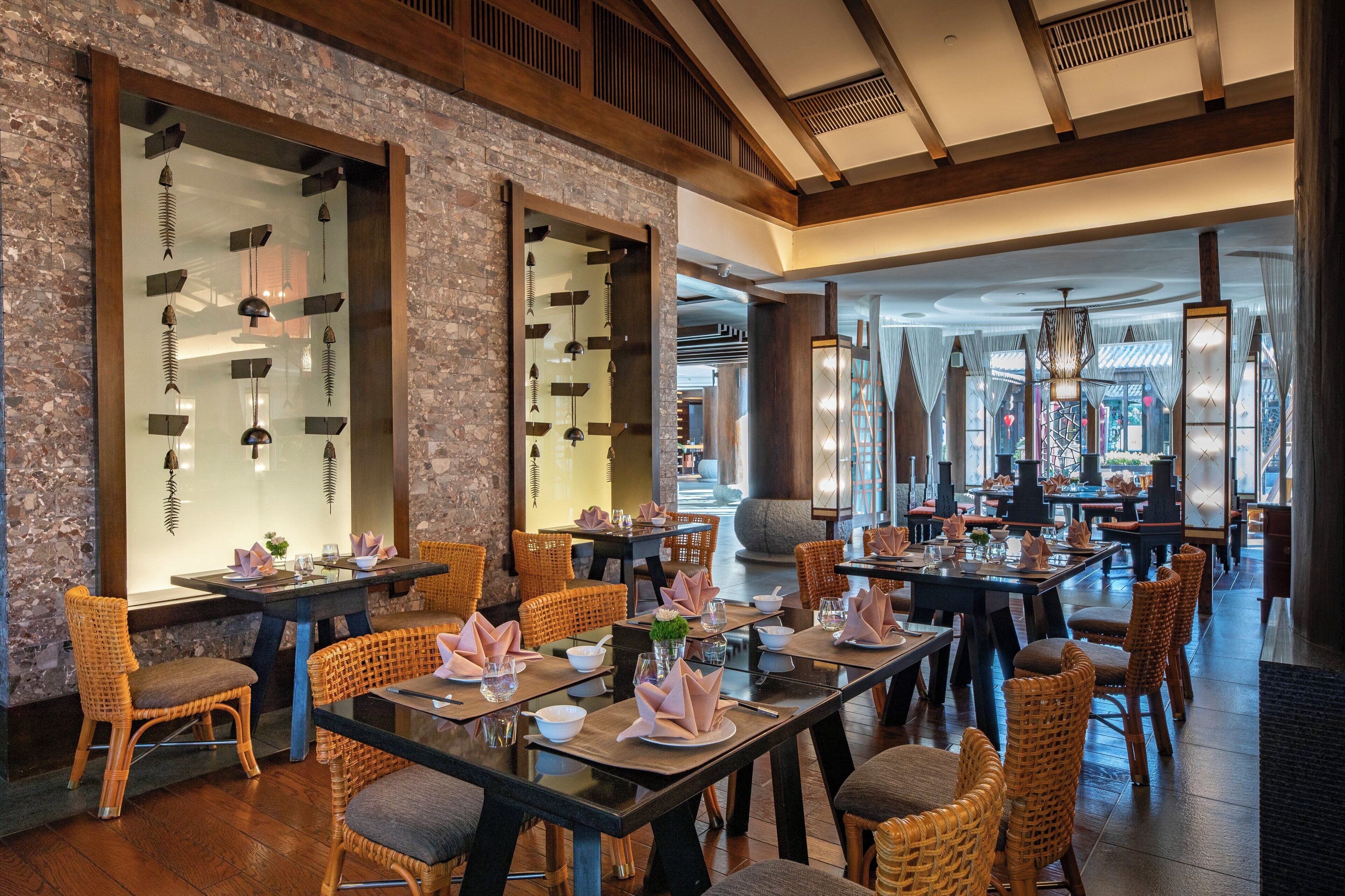 A restaurant with wooden tables and wicker chairs, large windows, and hanging light fixtures.