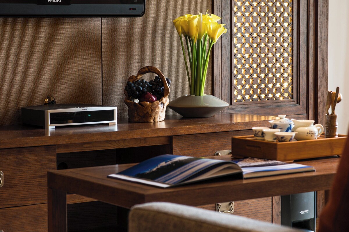 A wooden desk with a flat-screen TV, a vase with yellow flowers, a fruit bowl, and a tray with teacups.
