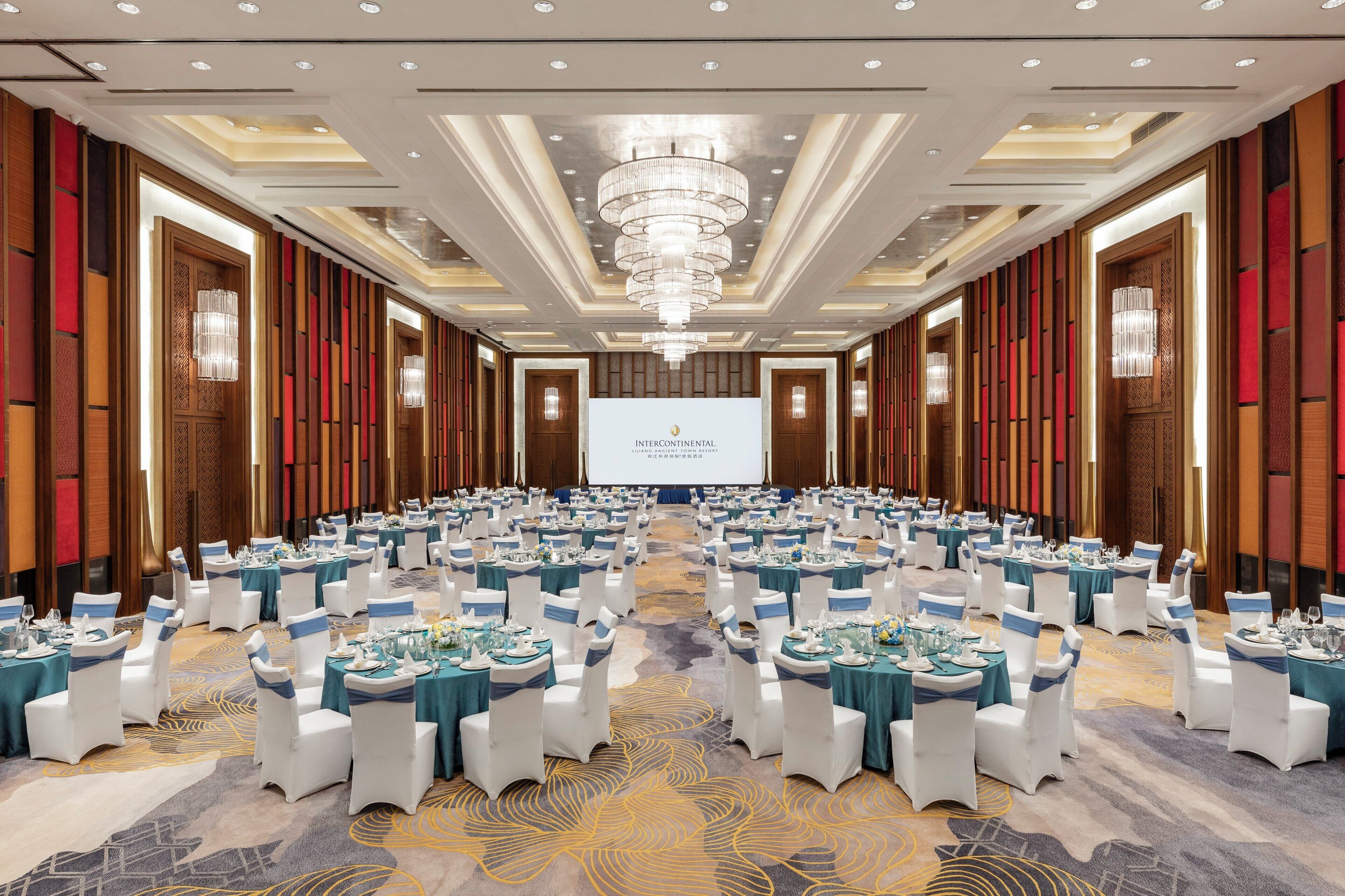 Una grande sala da banchetto con tavoli rotondi apparecchiati per un evento formale, con un palco centrale e un grande schermo.