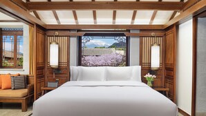 Una cama grande con sábanas blancas, cabecera de madera y mesitas de noche con lámparas. Se ve un banco de madera y una pequeña mesa con un jarrón de flores.
