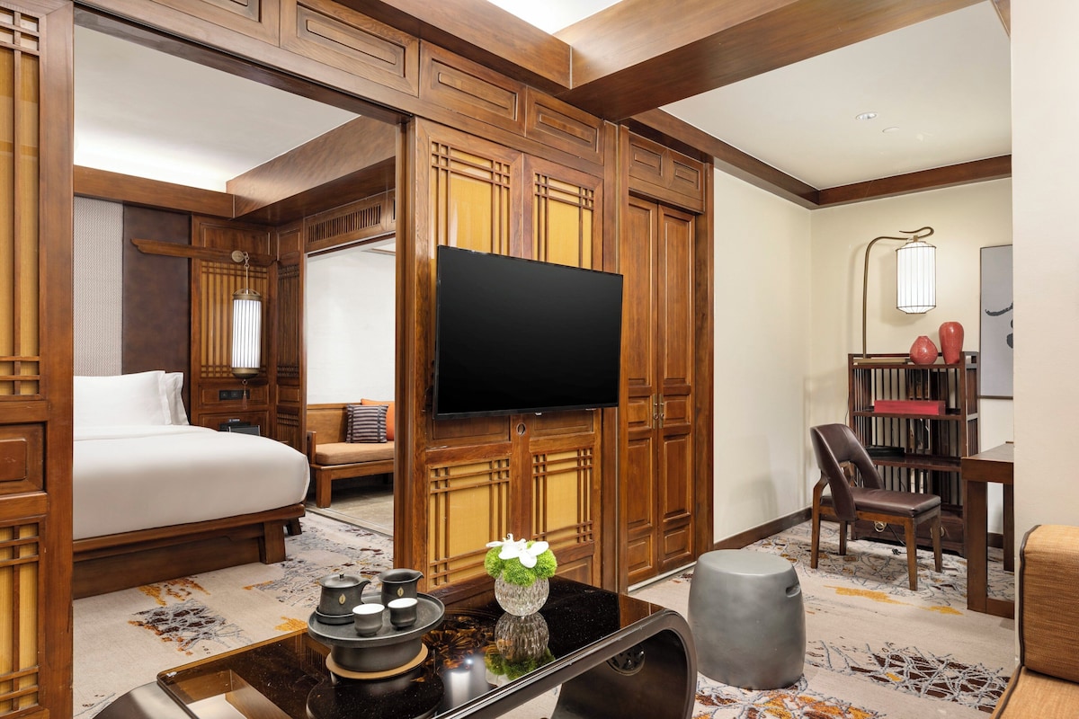 Una habitación de hotel con una cama, un televisor, un escritorio con un florero y una estantería.