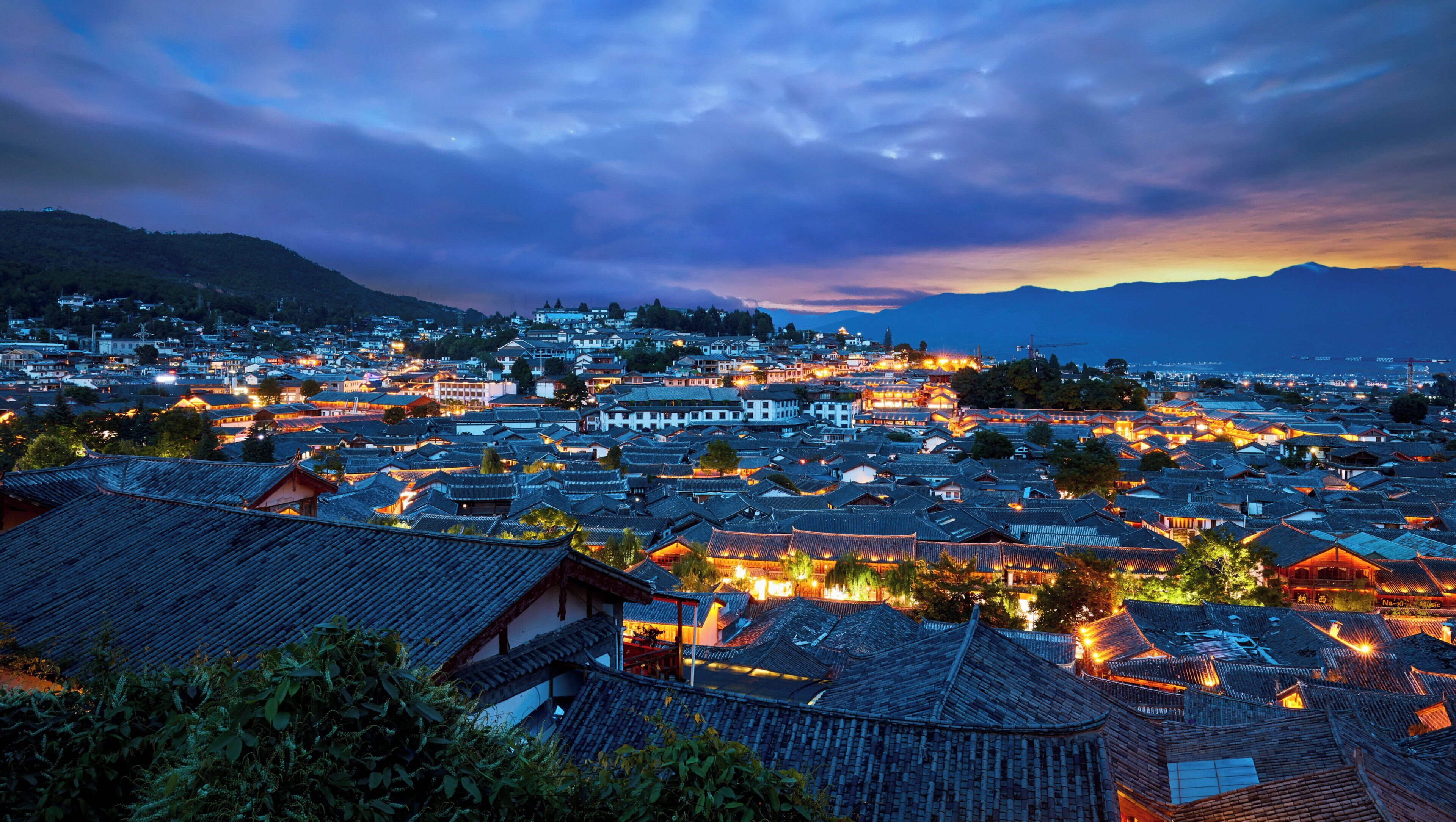 A traditional town with numerous buildings, some with illuminated signs, and a mountainous backdrop.