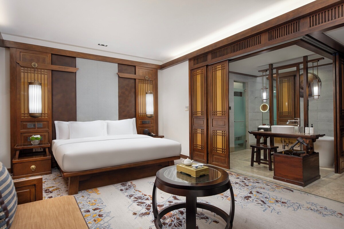 A hotel room with a large bed, a small round table, a wooden wardrobe, and a bathroom visible through a glass door.