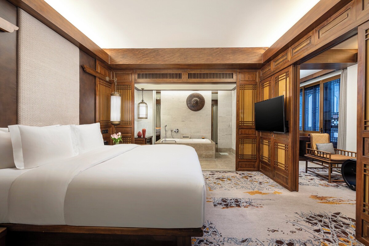 A spacious hotel room with a large bed, a television, a bathroom with a bathtub, and a view of the outside.