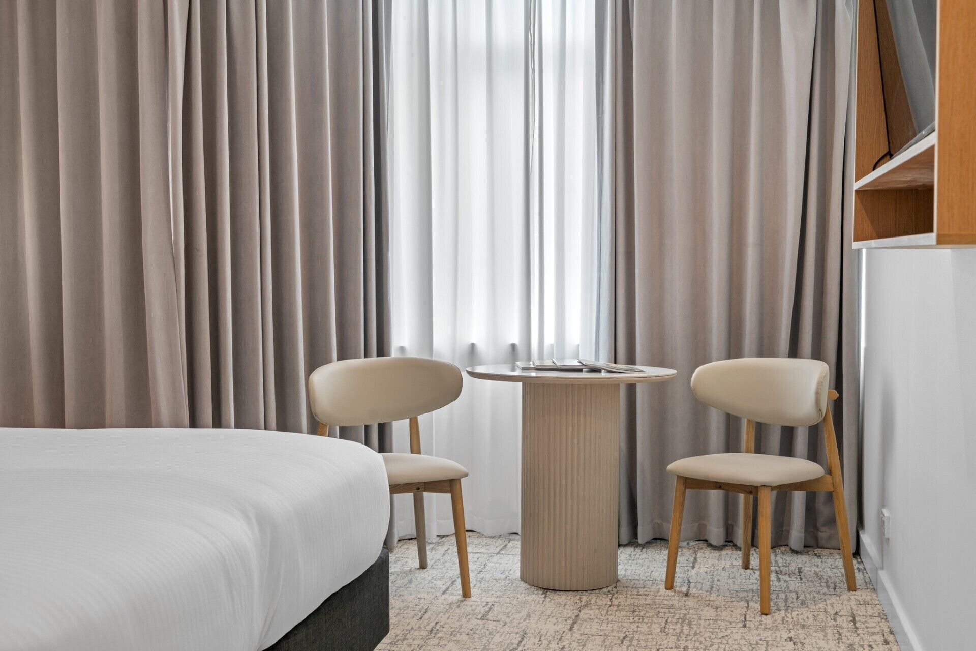 Une chambre d’hôtel avec un lit, deux chaises, une table ronde et une grande fenêtre avec des rideaux.