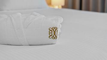 Une serviette blanche, soigneusement pliée, ornée d’un monogramme doré brodé, est posée sur le lit.