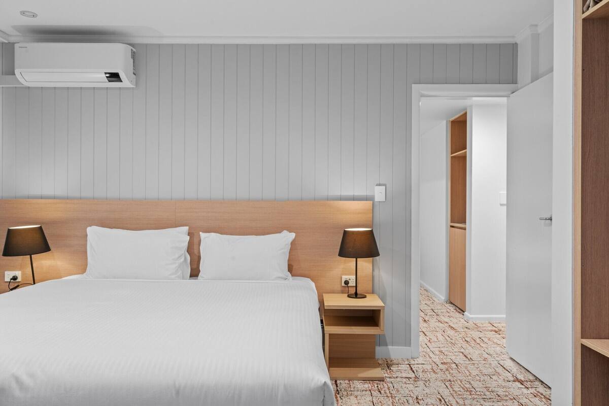 A hotel room with a bed, bedside lamps, a nightstand, a wall-mounted air conditioner, and a wooden wardrobe.