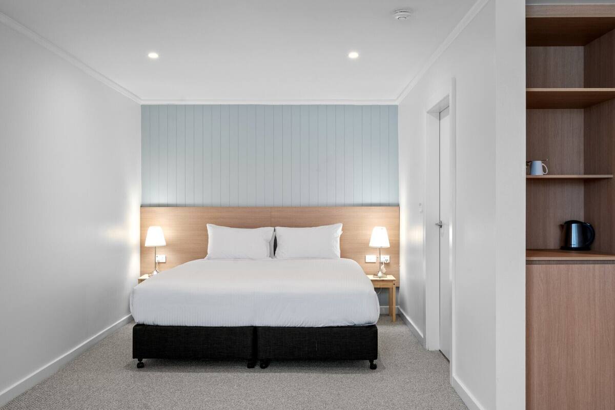 A modern hotel room with a large bed, two bedside lamps, a wooden headboard, and a built-in shelf with a kettle.