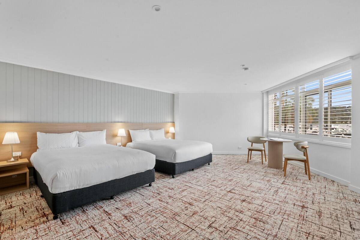 A hotel room with two beds, a desk, and a window with blinds.