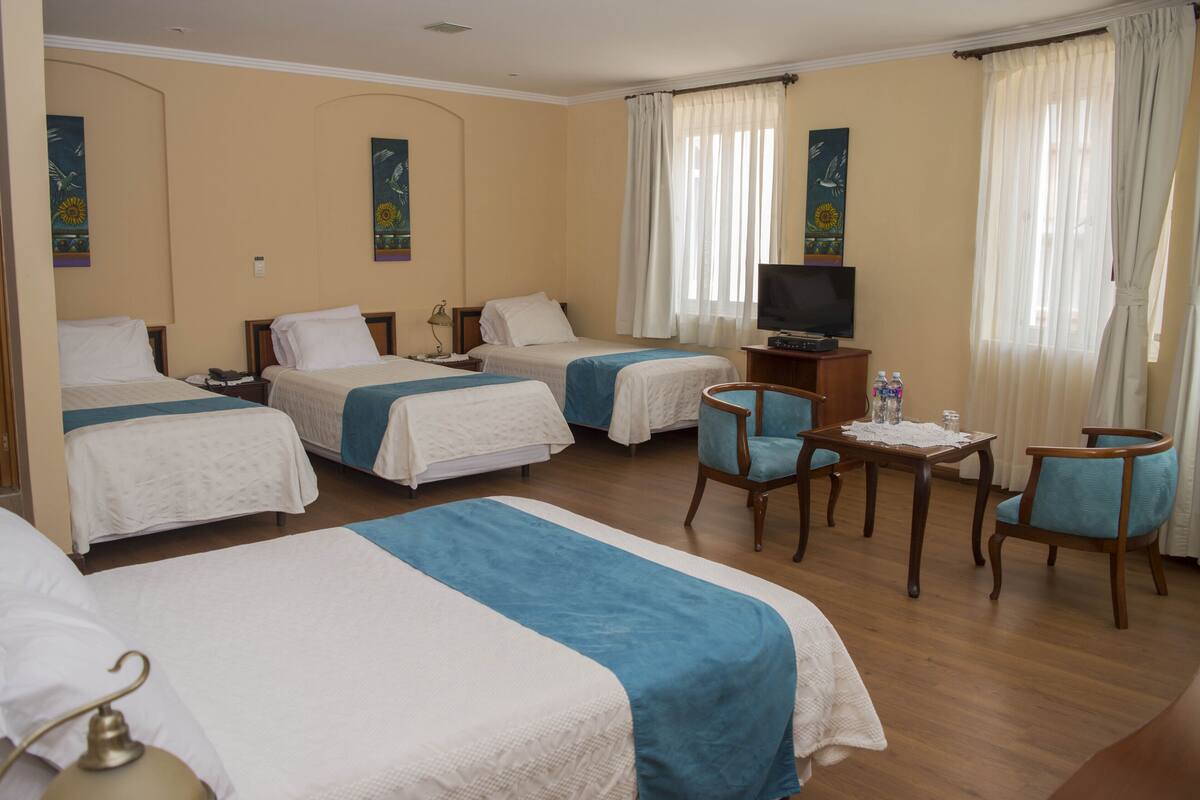 Une chambre d’hôtel avec trois lits, une télévision, une petite table avec une bouteille d’eau et deux chaises.