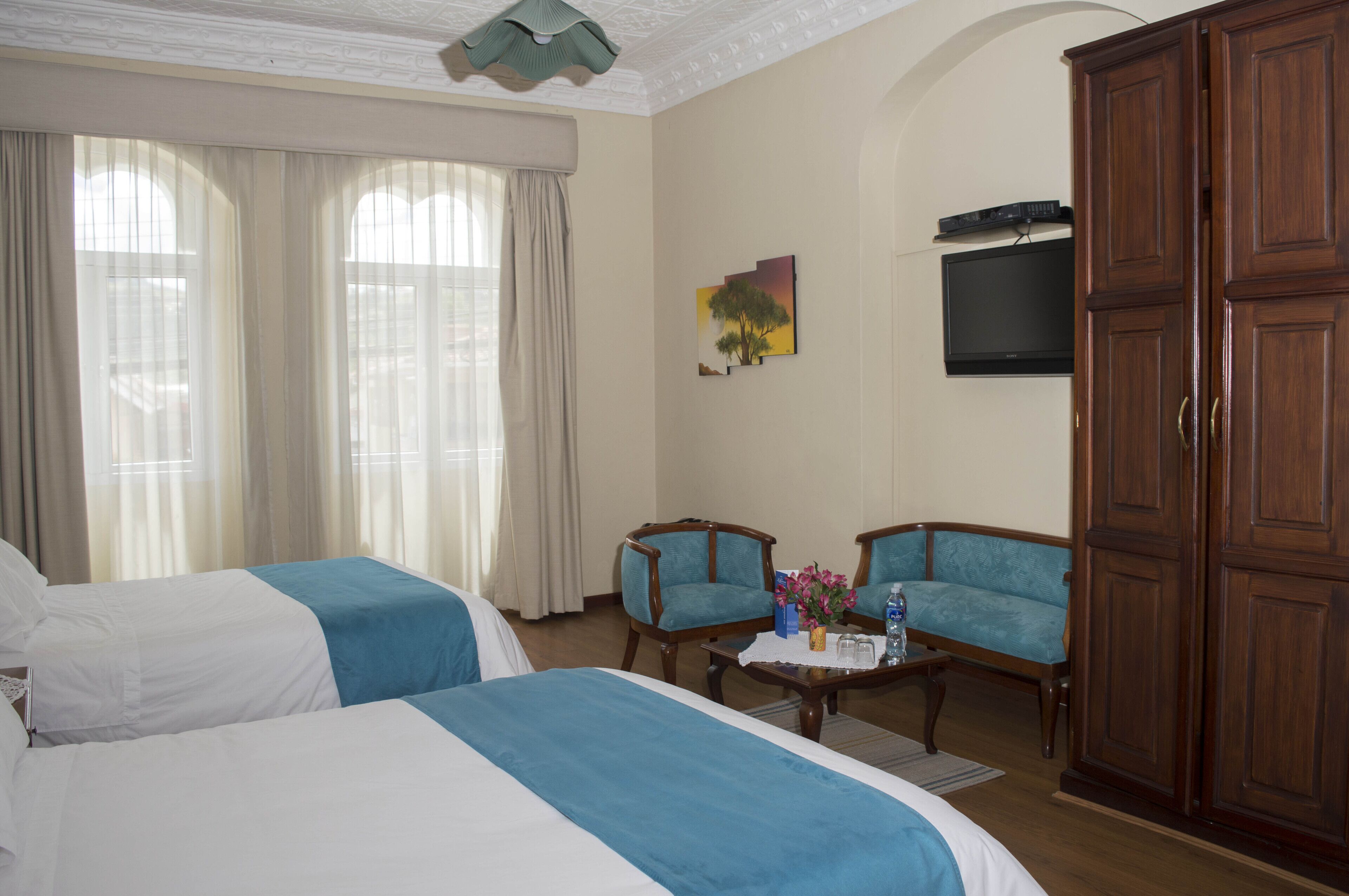 A hotel room with two beds, a television, a wooden wardrobe, and a small table with a vase of flowers.