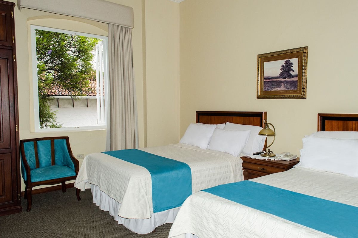 Une chambre d’hôtel avec deux lits, une chaise, une table de chevet avec une lampe, un tableau encadré au mur et une fenêtre avec des rideaux.