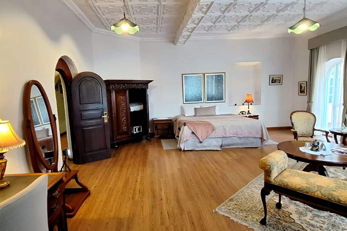 Une chambre d’hôtel comprenant un lit, un bureau avec une lampe, une chaise, un canapé, une armoire en bois et une grande fenêtre avec des rideaux.