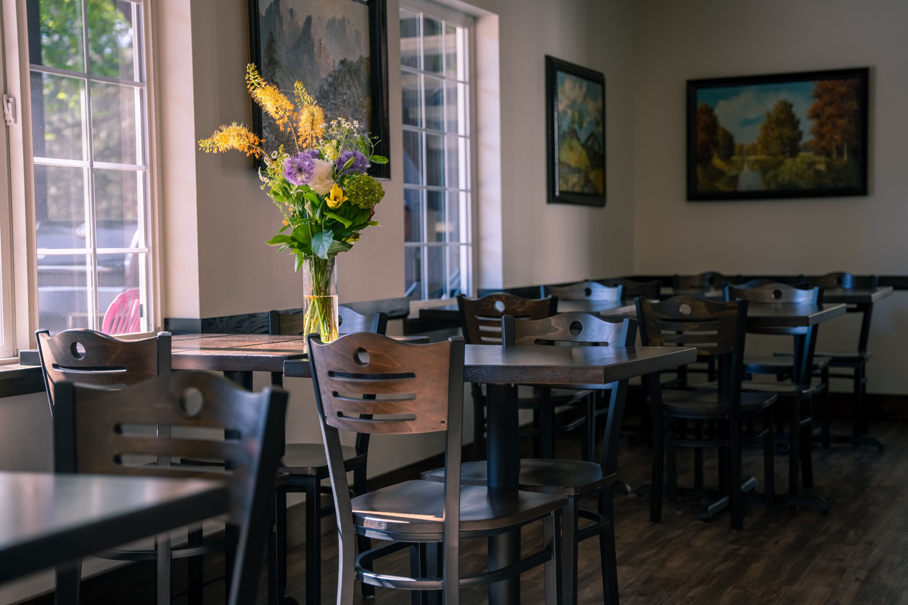 A restaurant interior with wooden tables and chairs, a vase of flowers on the table, and paintings on the walls.