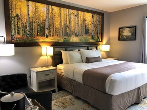 A hotel room with a large forest painting above the bed, two bedside lamps, a black leather chair, a small bedside table with a lamp, and a patterned rug. - Blue Elk Inn (Leavenworth)