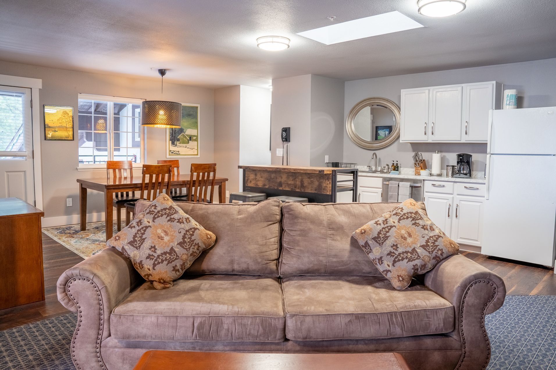 A cozy living room with a sofa, a dining table, and chairs. There is a kitchen area with cabinets and a refrigerator.