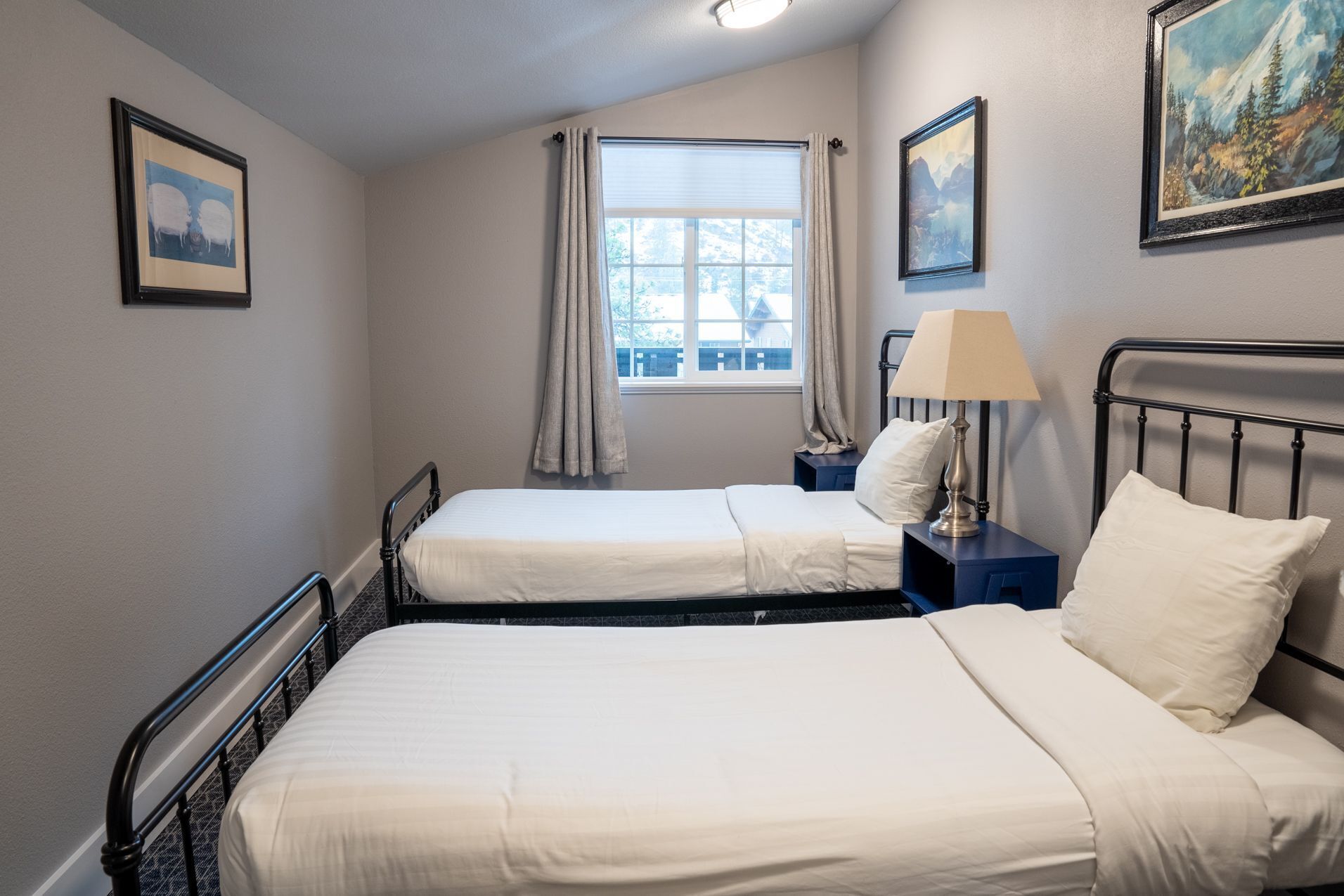 Habitación doble con dos camas, una ventana con cortinas, una mesita de noche con lámpara y cuadros enmarcados en las paredes.