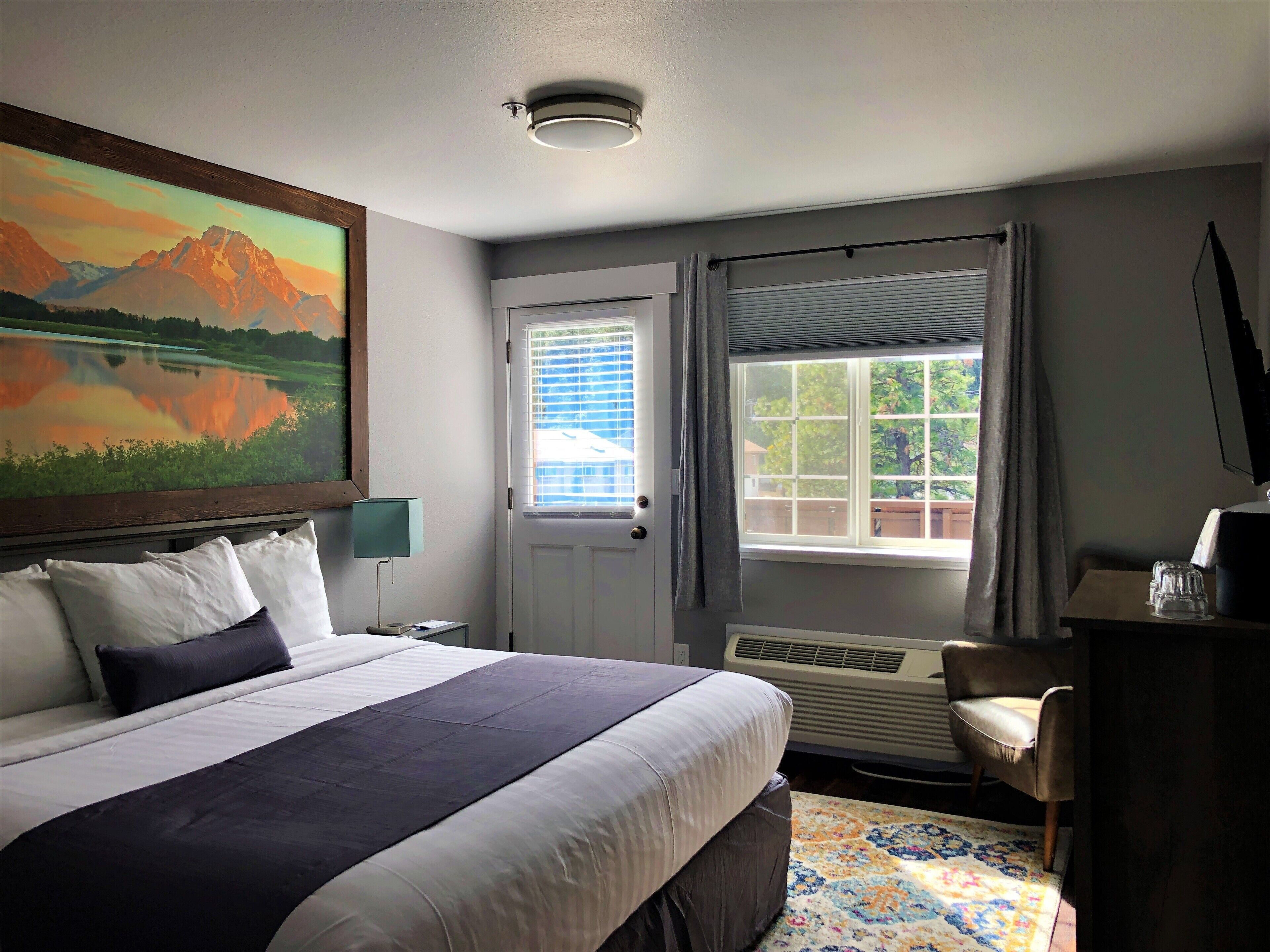 A hotel room with a large bed, a painting of mountains, a window with curtains, a desk with a lamp, and a chair.