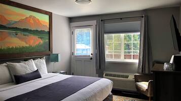 Quarto de hotel com uma cama grande, um quadro de montanhas, uma janela com cortinas, uma escrivaninha com abajur e uma cadeira.