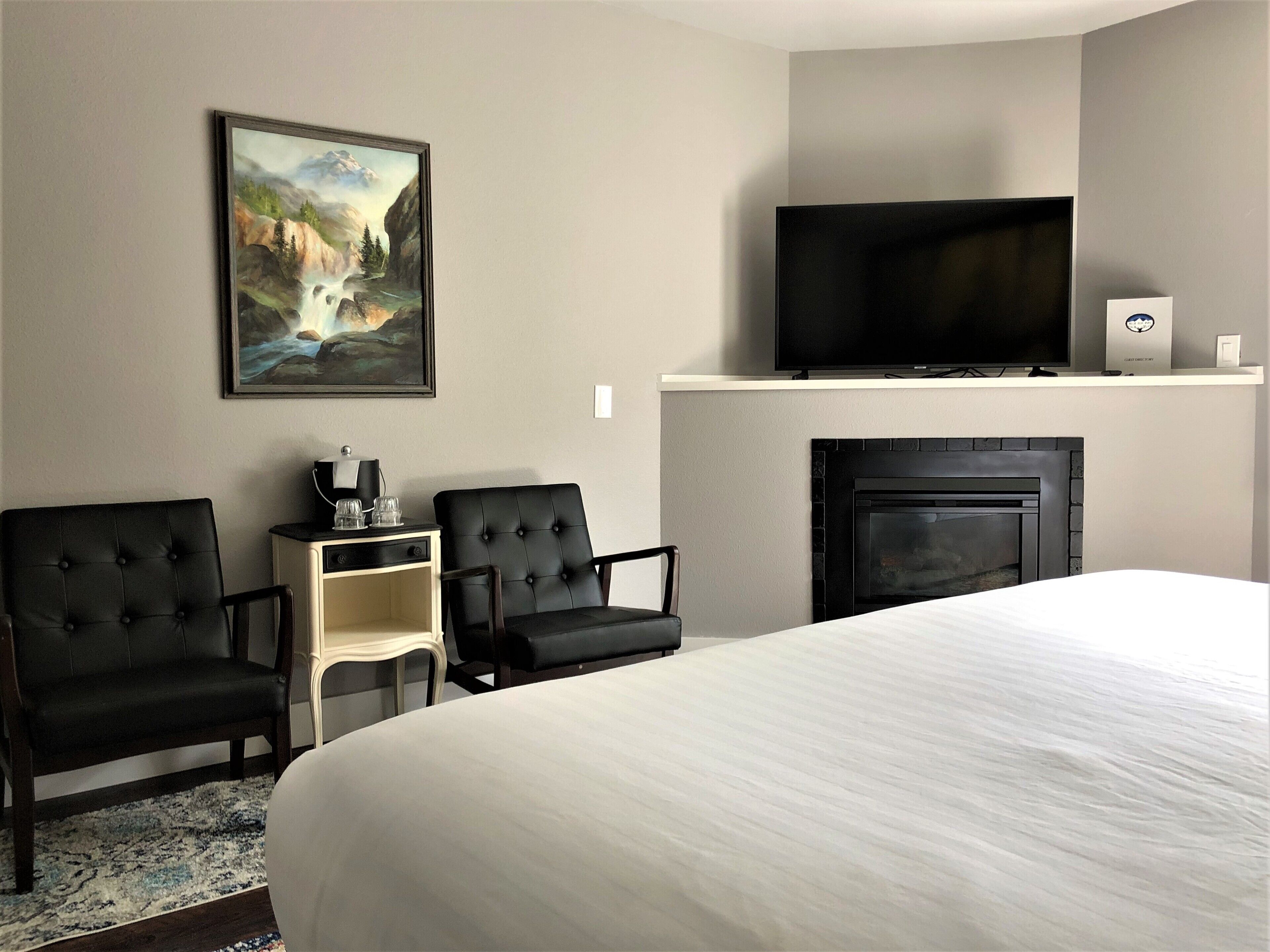 A hotel room with a large bed, two black armchairs, a small side table with a lamp, a flat-screen TV on a shelf above a fireplace, and a framed painting on the wall.