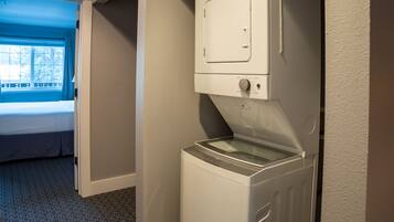 A compact laundry room with a front-loading washing machine and a dryer, a small window with curtains, and a bed in the adjacent room.