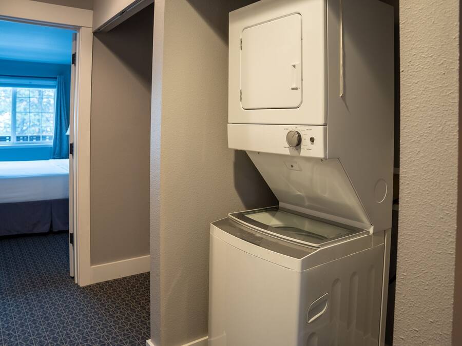 A compact laundry room with a front-loading washing machine and a dryer, a small window with curtains, and a bed in the adjacent room.