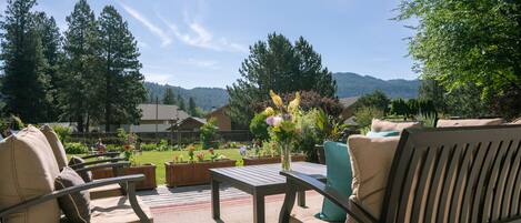 Een terras buiten met een bank, tafel en stoelen. Op de tafel staat een vaas met bloemen. Het terras heeft uitzicht op een tuin met diverse planten en een bergzicht op de achtergrond.