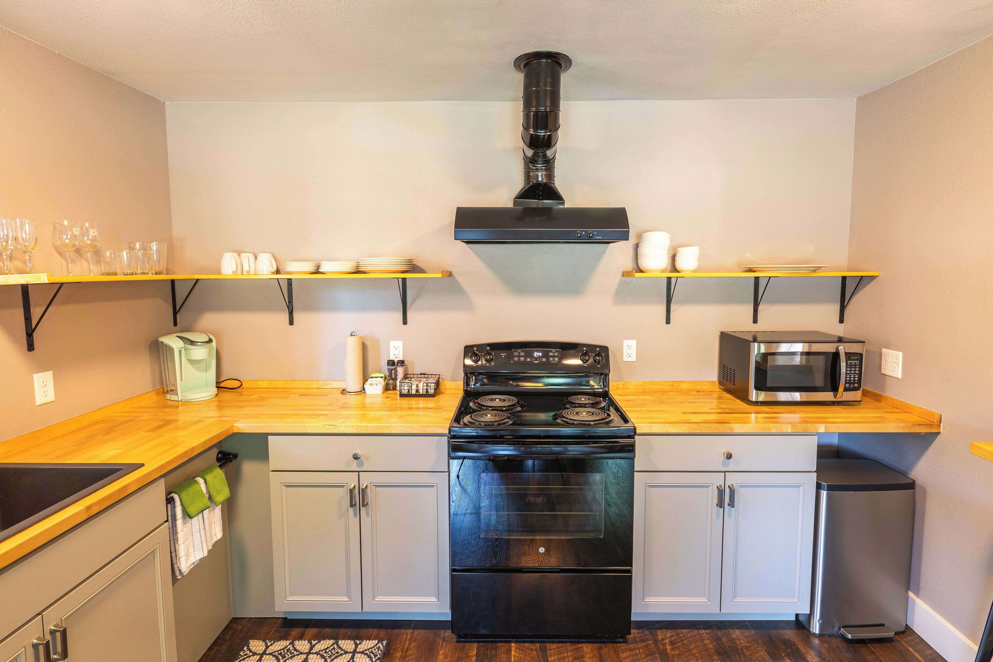 A compact kitchen with a black stove, a microwave, and a sink. There are wooden shelves above the stove holding glasses and cups.