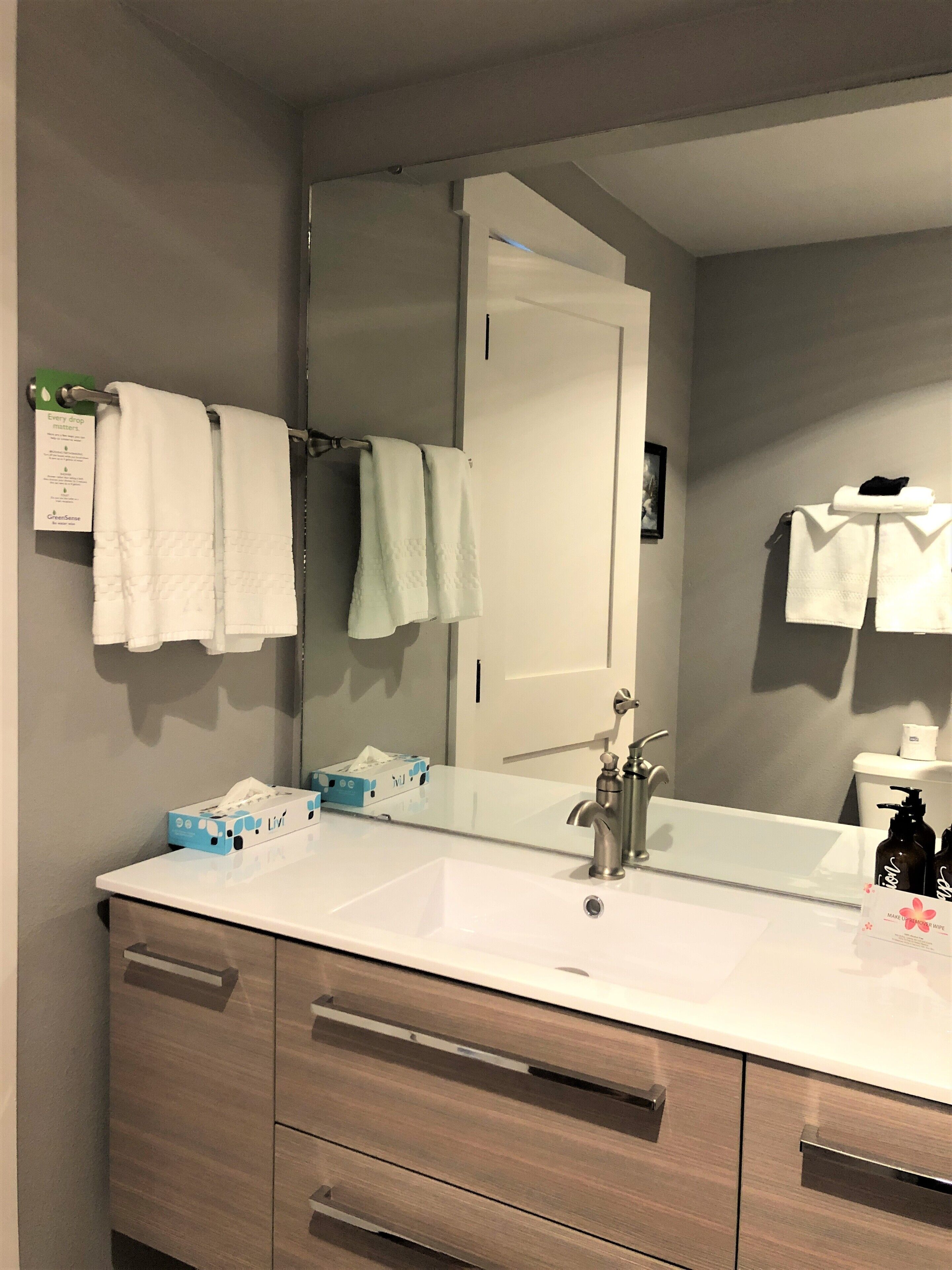 A bathroom with a white sink, a mirror, and two towels hanging on a rack. There are toiletries on the sink and a tissue box on the counter.