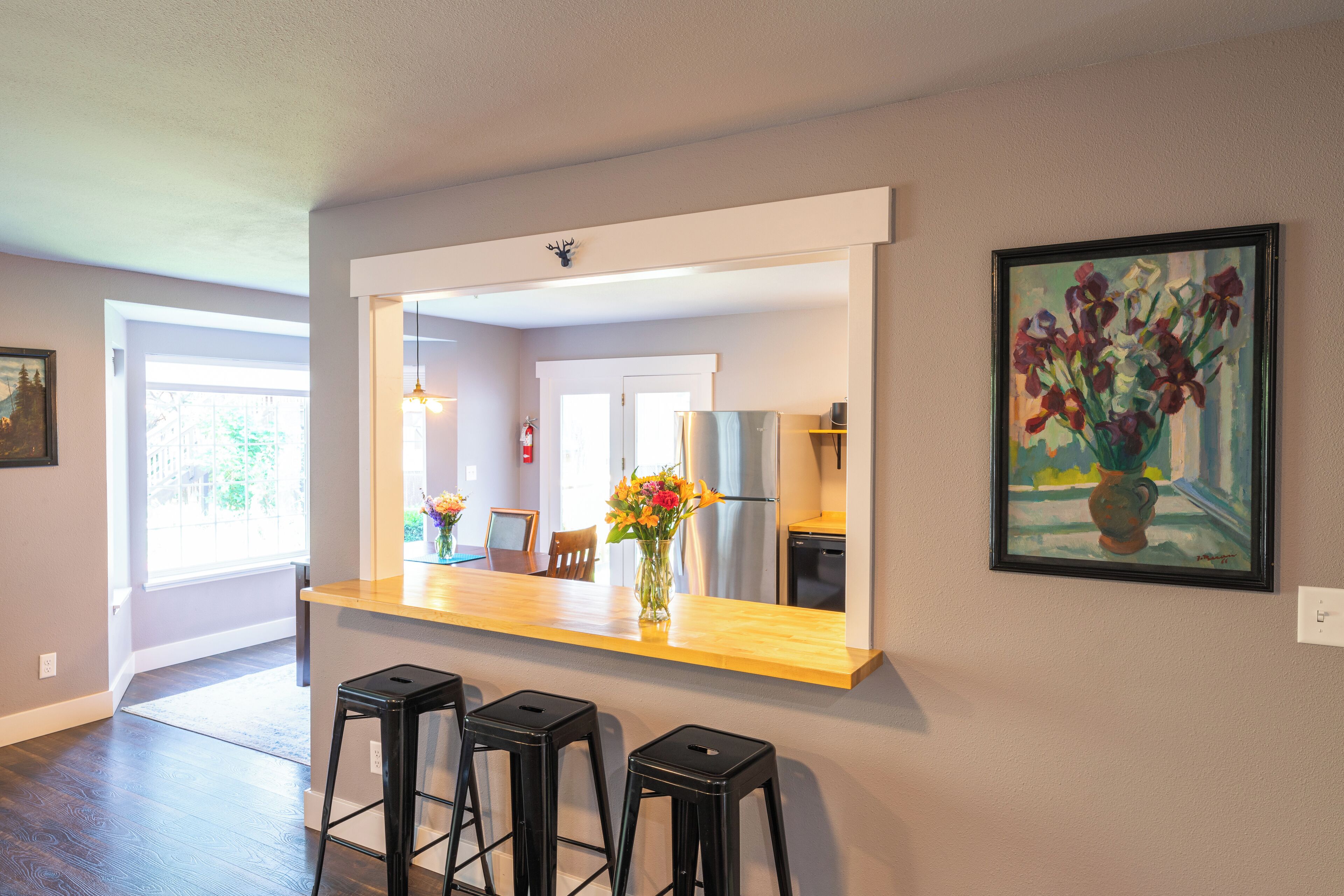 Une cuisine avec un plan de travail en bois, trois tabourets noirs, un vase de fleurs et un tableau encadré accroché au mur.