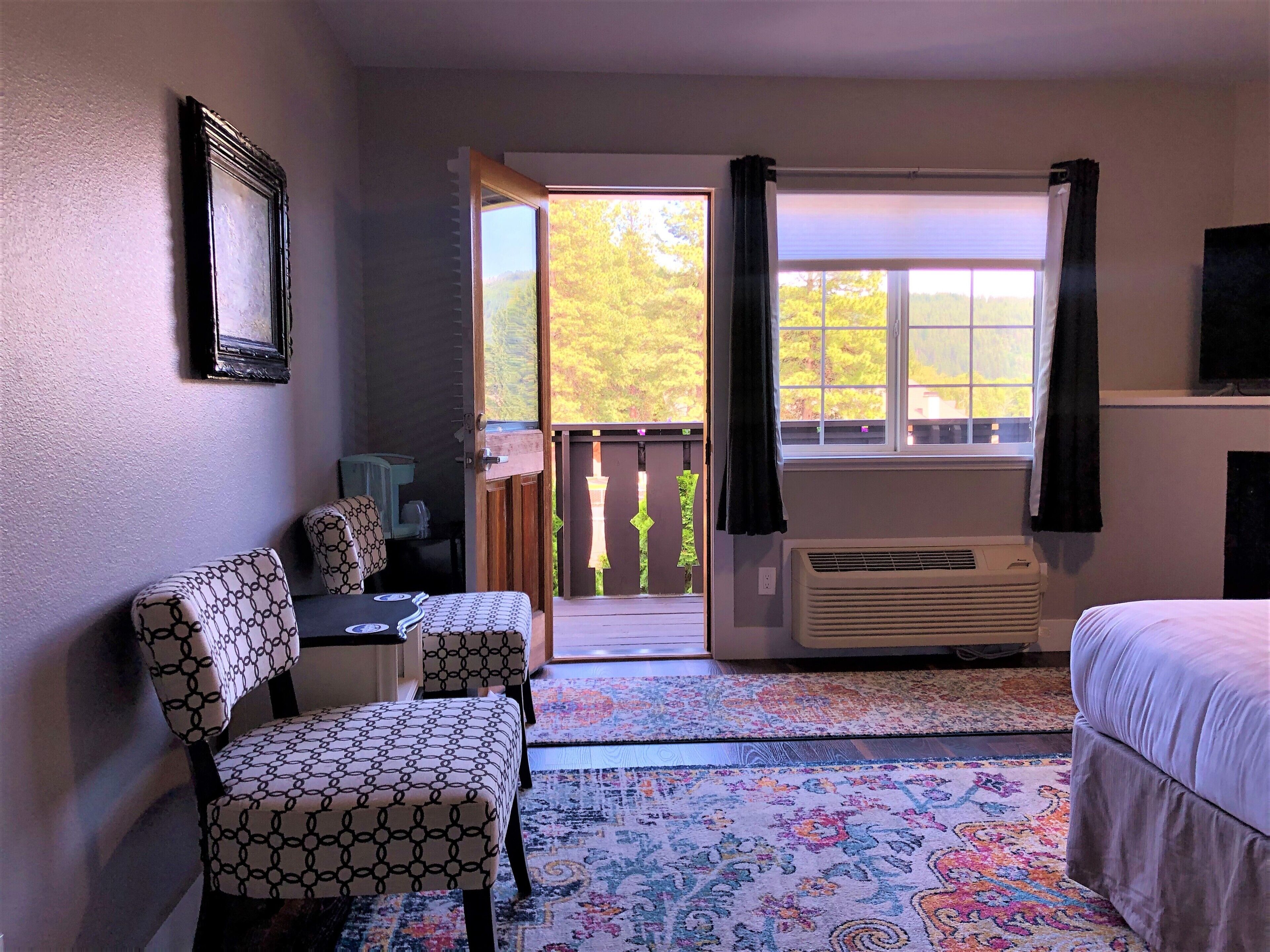A hotel room with a bed, two patterned chairs, a small table, a window with curtains, an air conditioner, and a view of trees.