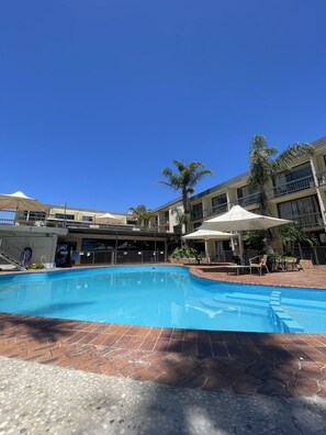A swimming pool with a tiled deck, surrounded by palm trees and a building with balconies. - El Lago Waters Motel (The Entrance)