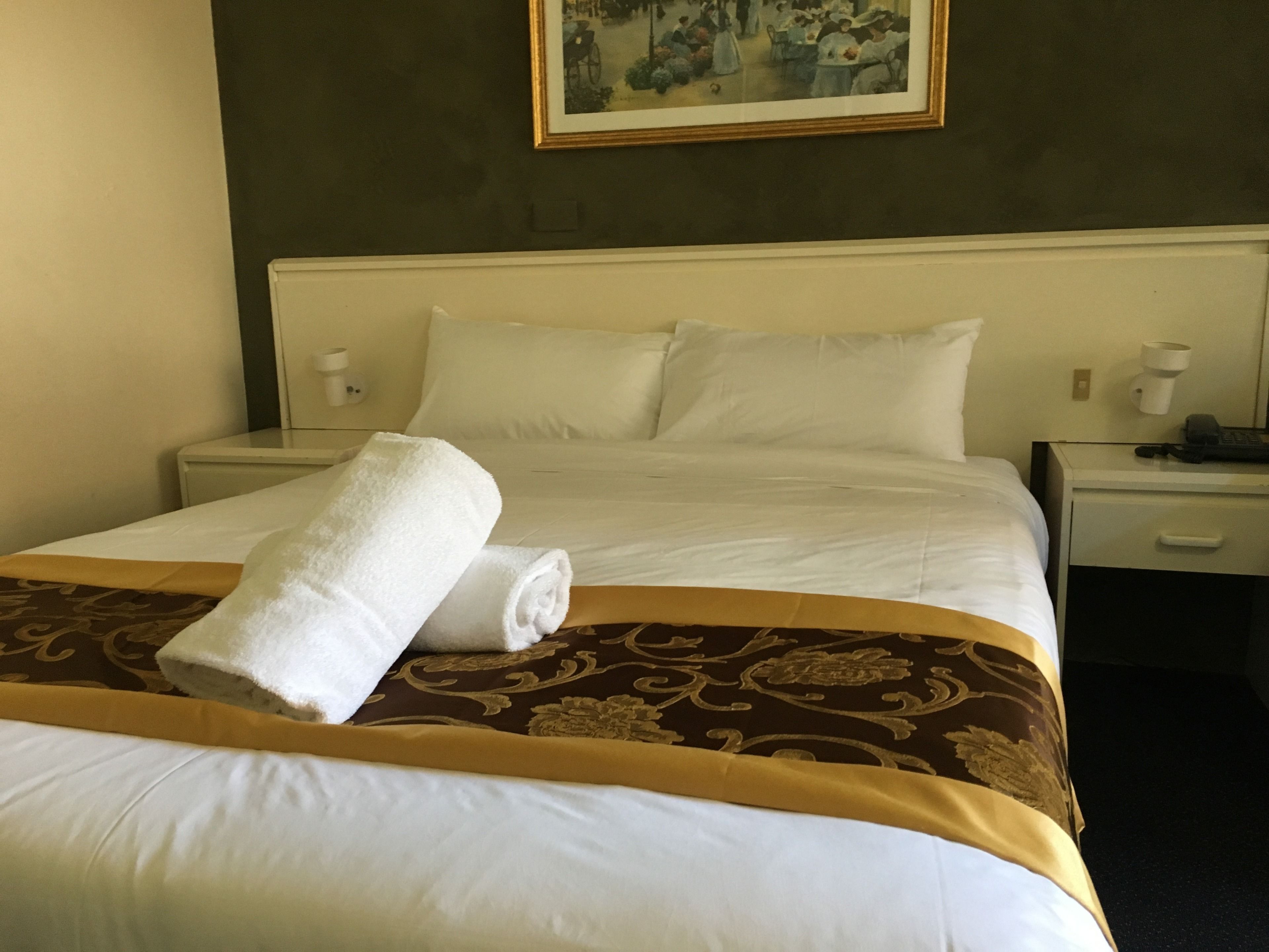 A neatly made bed with a rolled white towel, a brown patterned bedspread, and a painting above the headboard.