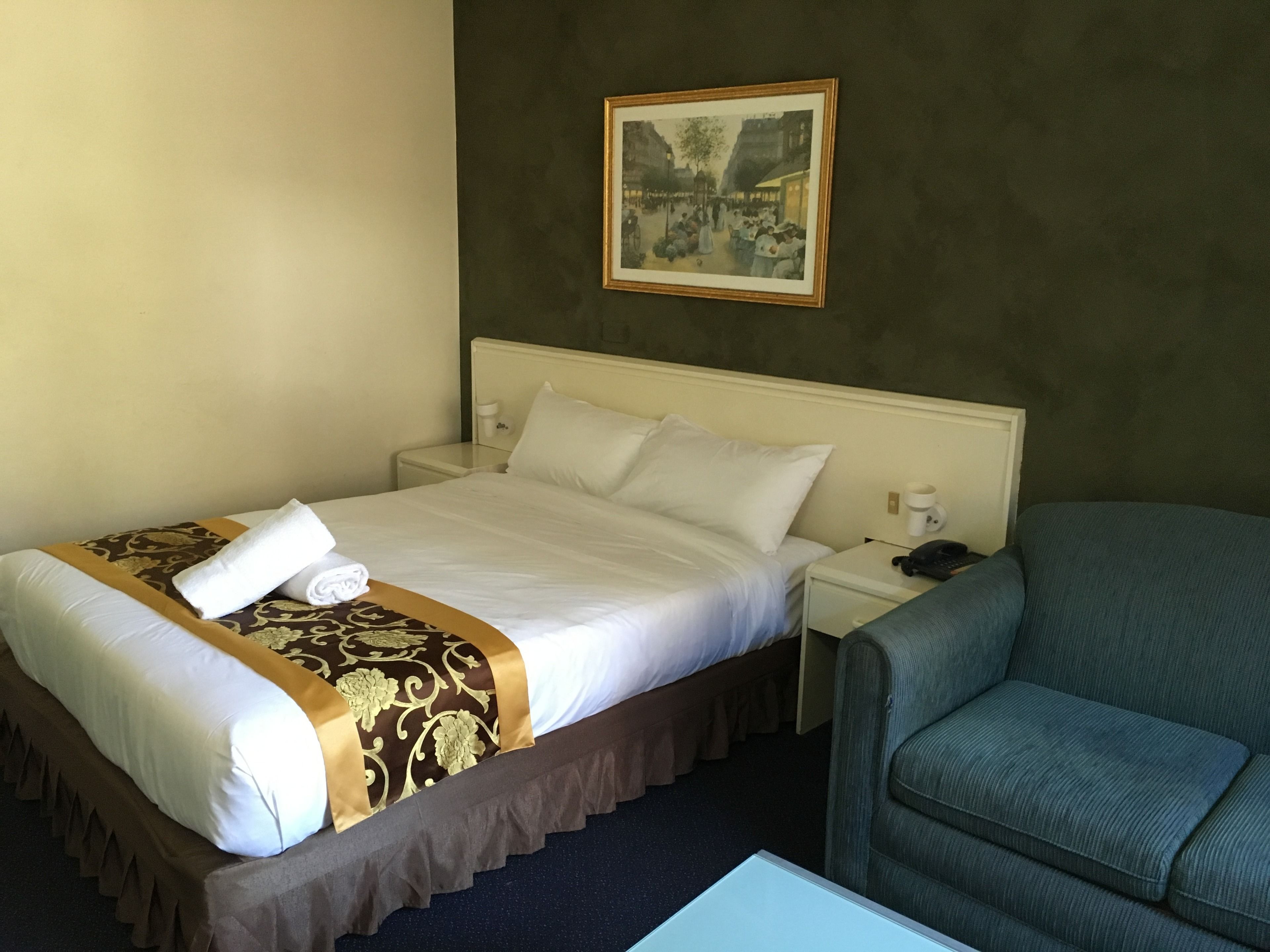 A hotel room with a neatly made bed, a chair, a bedside table with a lamp, and a framed picture on the wall.
