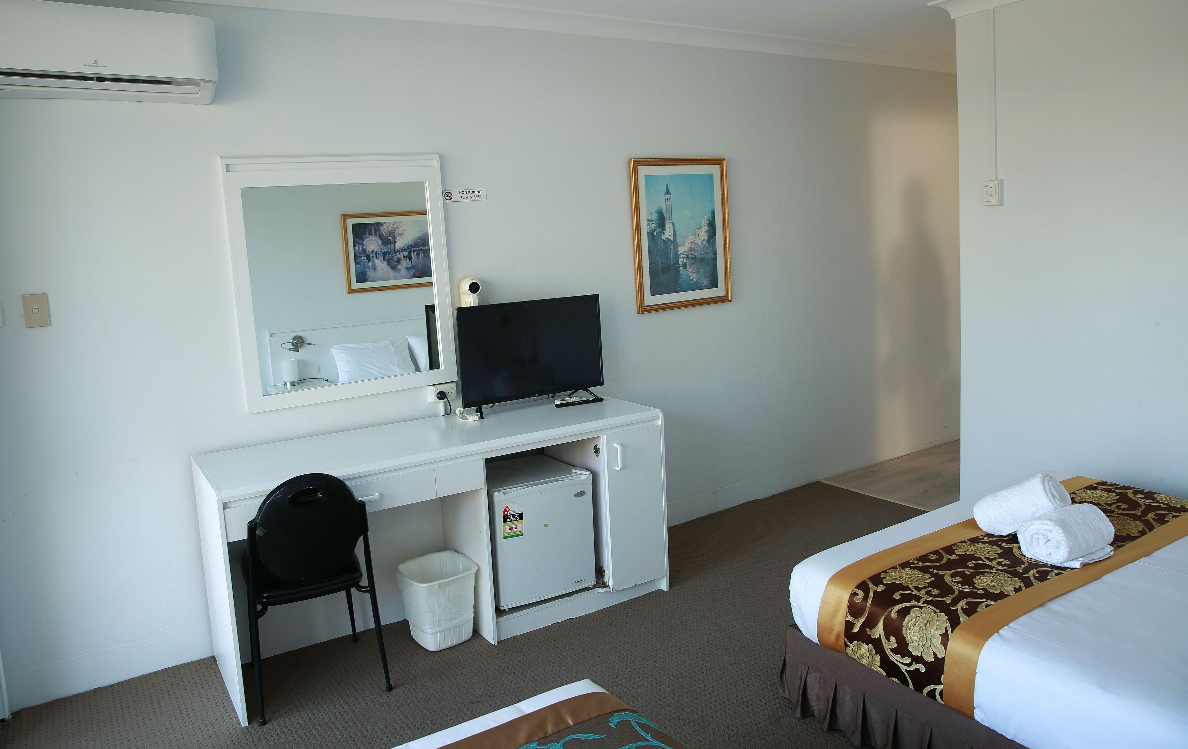 A hotel room with a white desk, a television, a small refrigerator, a chair, a bed with a decorative bedspread, and two framed pictures on the wall.