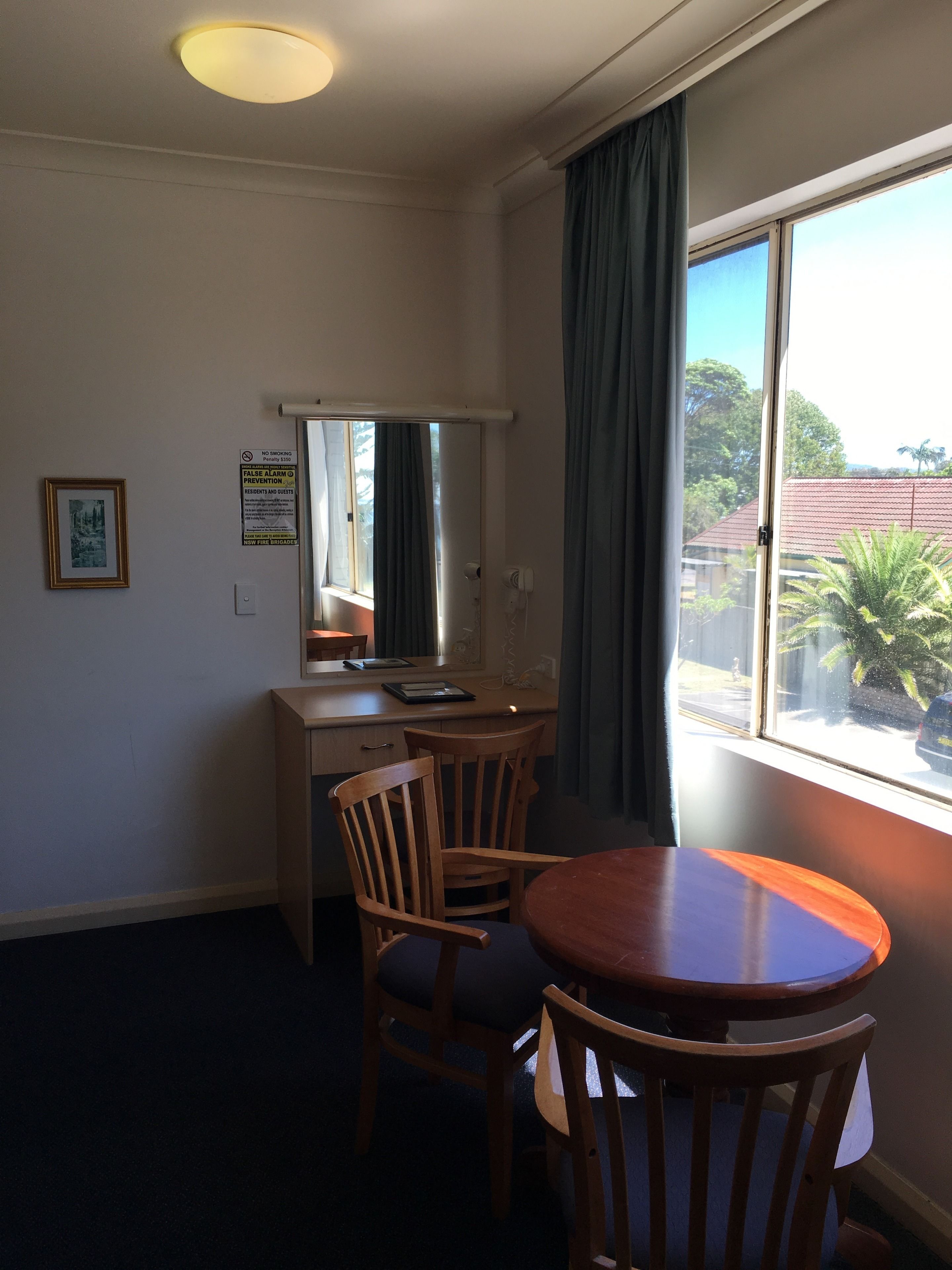 A room with a desk, two chairs, a round table, a window with curtains, a framed picture on the wall, and a ceiling light.