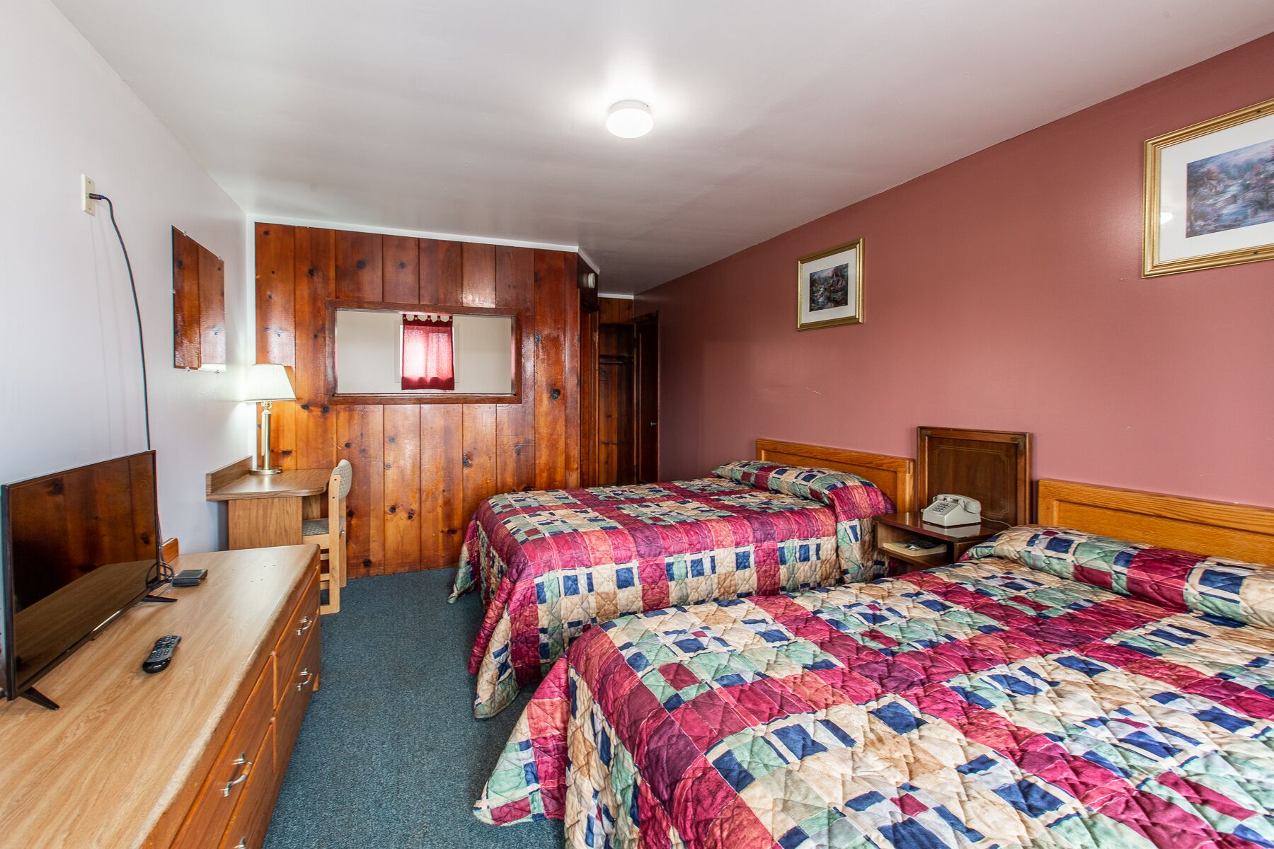 A bedroom with two beds, wooden walls, a desk, a lamp, and a framed picture on the wall.