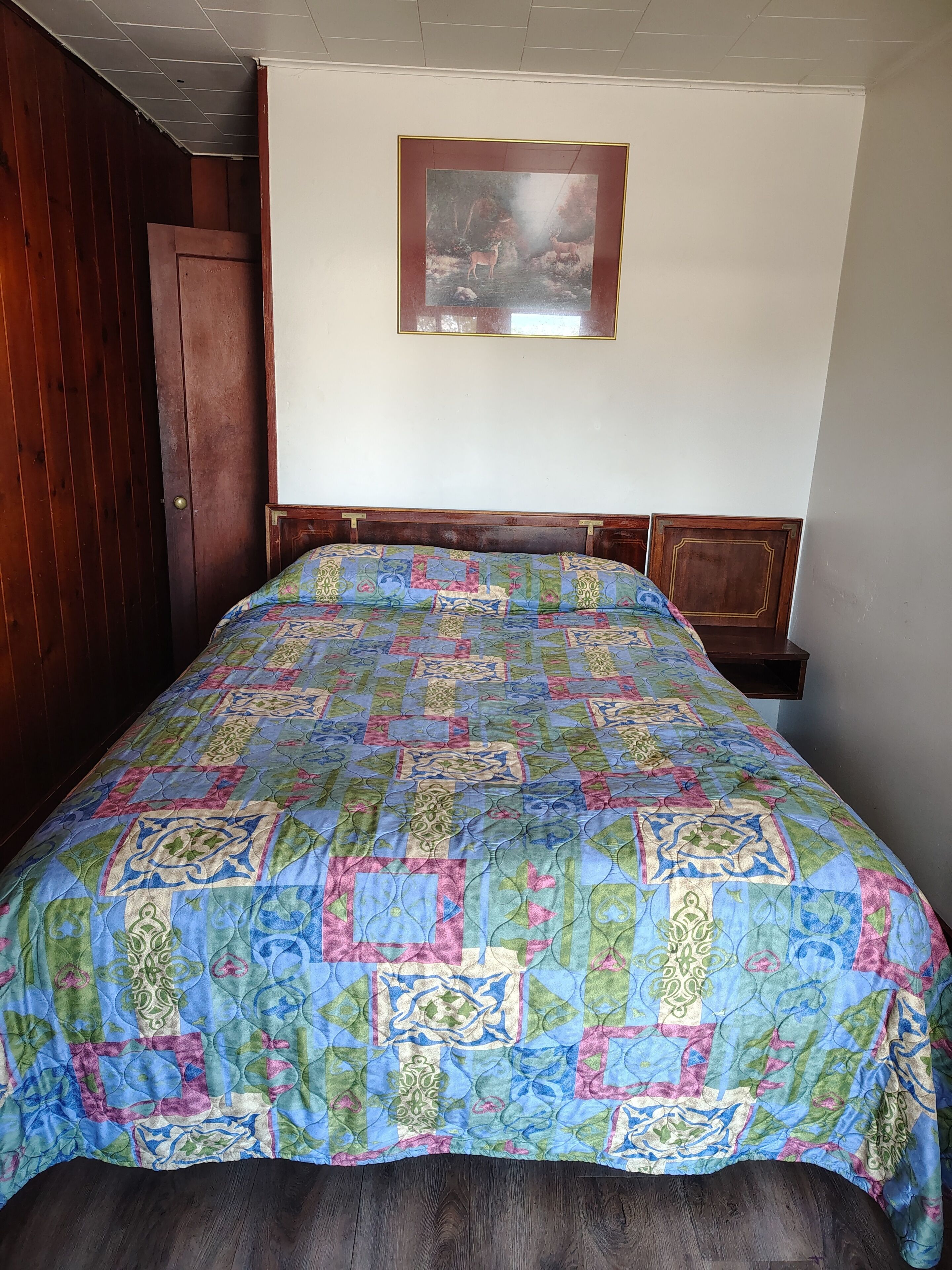 A bedroom with a wooden bed frame, a colorful quilt, a framed picture on the wall, and a wooden door.