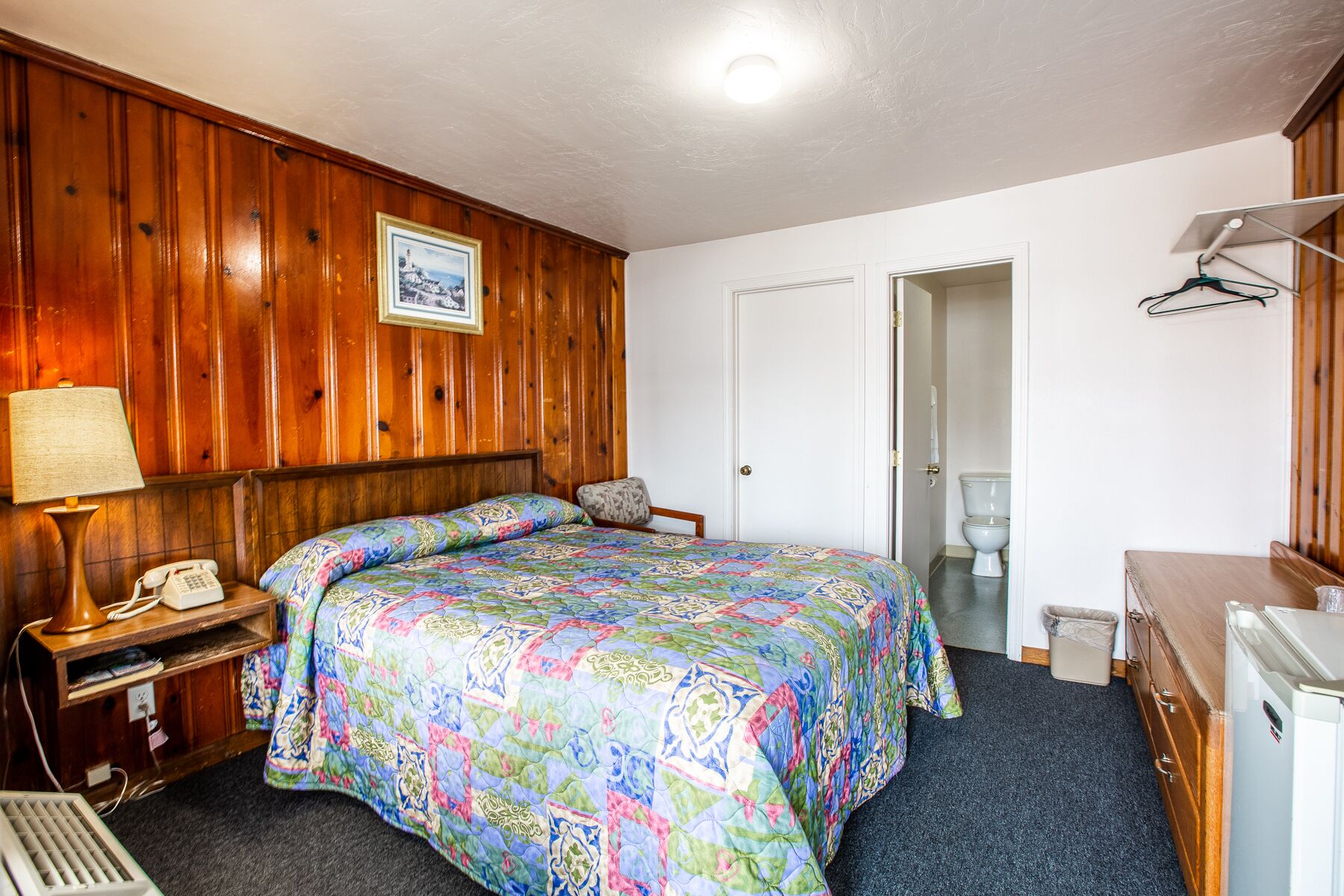 Una habitación con un muro revestido de paneles de madera, una cama con colcha de colores, un teléfono en la mesita de noche, una pequeña mesa con lámpara, una puerta que da al baño y un refrigerador.