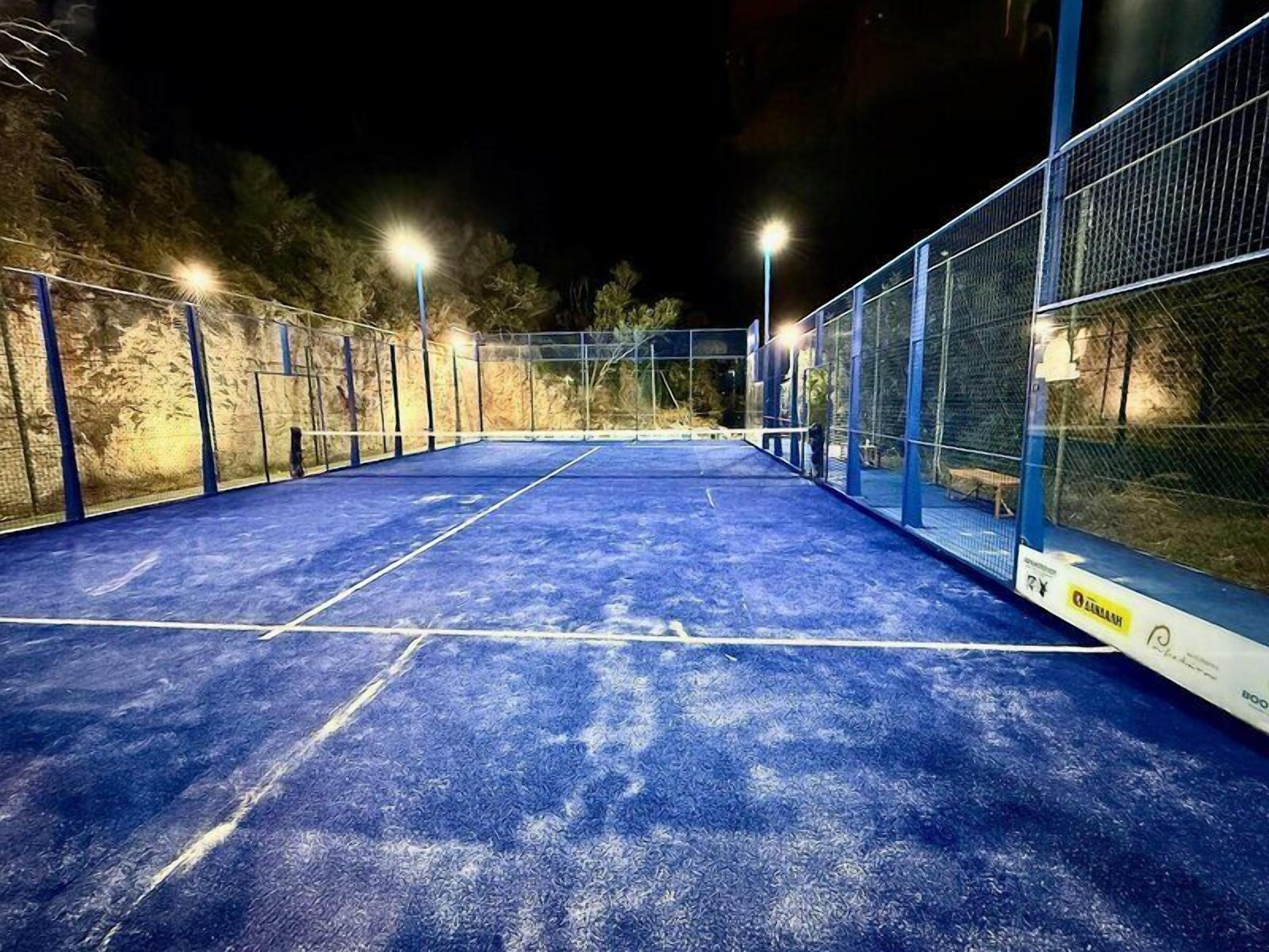 Campo da tennis all'aperto illuminato di notte, con pavimentazione e recinzione blu.
