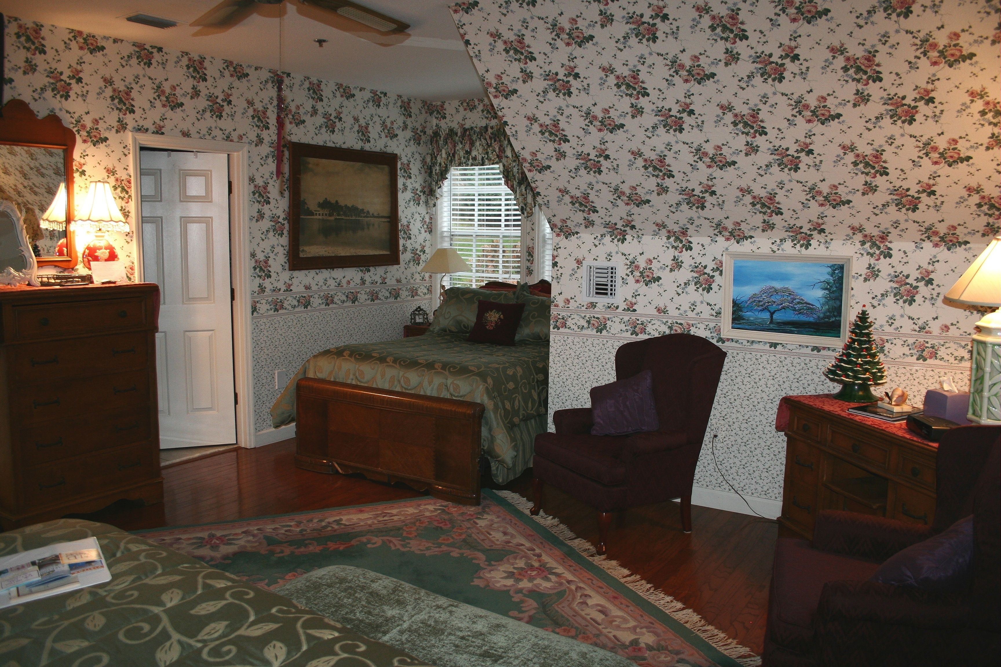 A bedroom with floral wallpaper, a bed, a chair, a dresser, a lamp, and a small Christmas tree.
