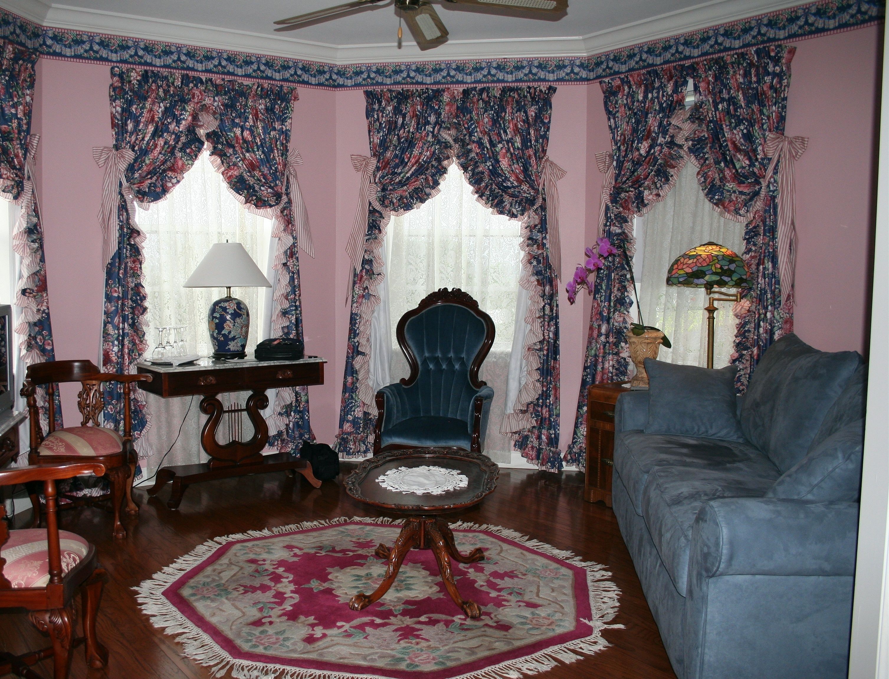 Una habitación con un sofá azul, una silla de madera, una mesita con una lámpara y cortinas con motivos florales.