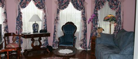 Una habitación con un sofá azul, una silla de madera, una mesita con una lámpara y cortinas con motivos florales.