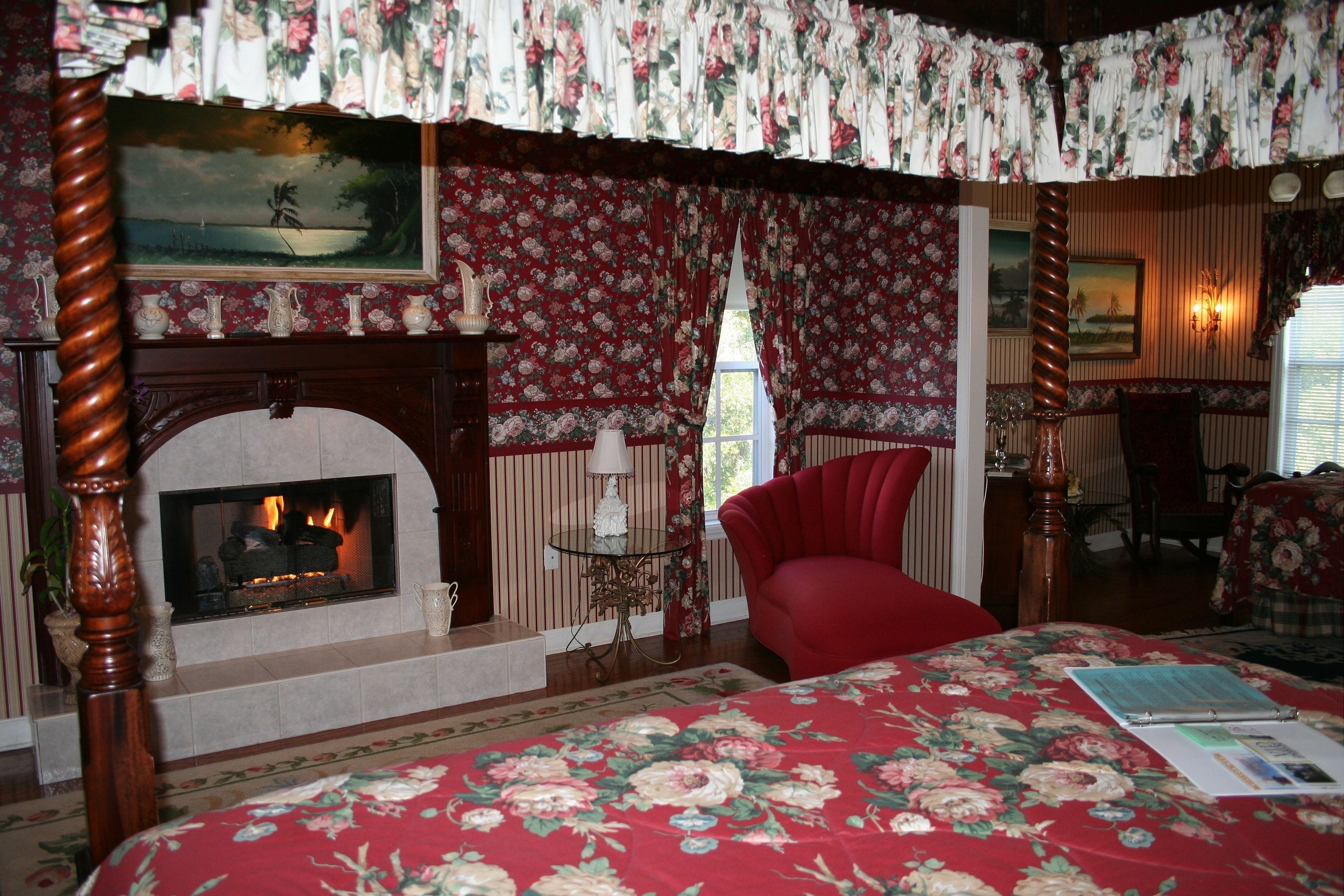 Un dormitorio con chimenea, papel tapiz floral, un sillón rojo y una cama con colcha estampada.