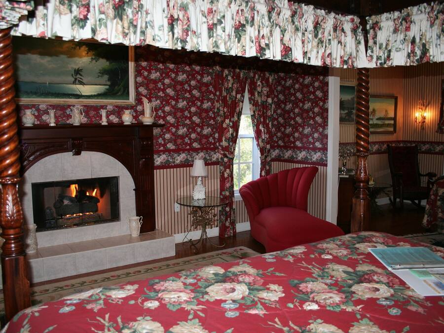 Un dormitorio con chimenea, papel tapiz floral, un sillón rojo y una cama con colcha estampada.